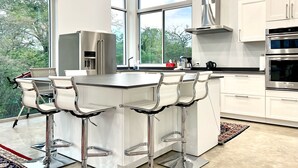 A modern kitchen with a central island, bar stools, stainless steel appliances, and large windows offering a view of greenery.