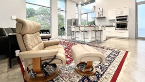A modern living room with a large window, a sofa, a coffee table, and a kitchen area with white cabinets and stainless steel appliances.