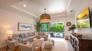 A modern living room with a gray sectional sofa, a round coffee table, wicker chairs, and a large TV on the wall. - Coastal Luxury Home with Pool, Spa & Gourmet Kitchen (Naples)