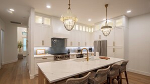 A modern kitchen with white cabinetry, a large island with bar stools, stainless steel appliances, and pendant lighting. - Coastal Luxury Home with Pool, Spa & Gourmet Kitchen (Naples)
