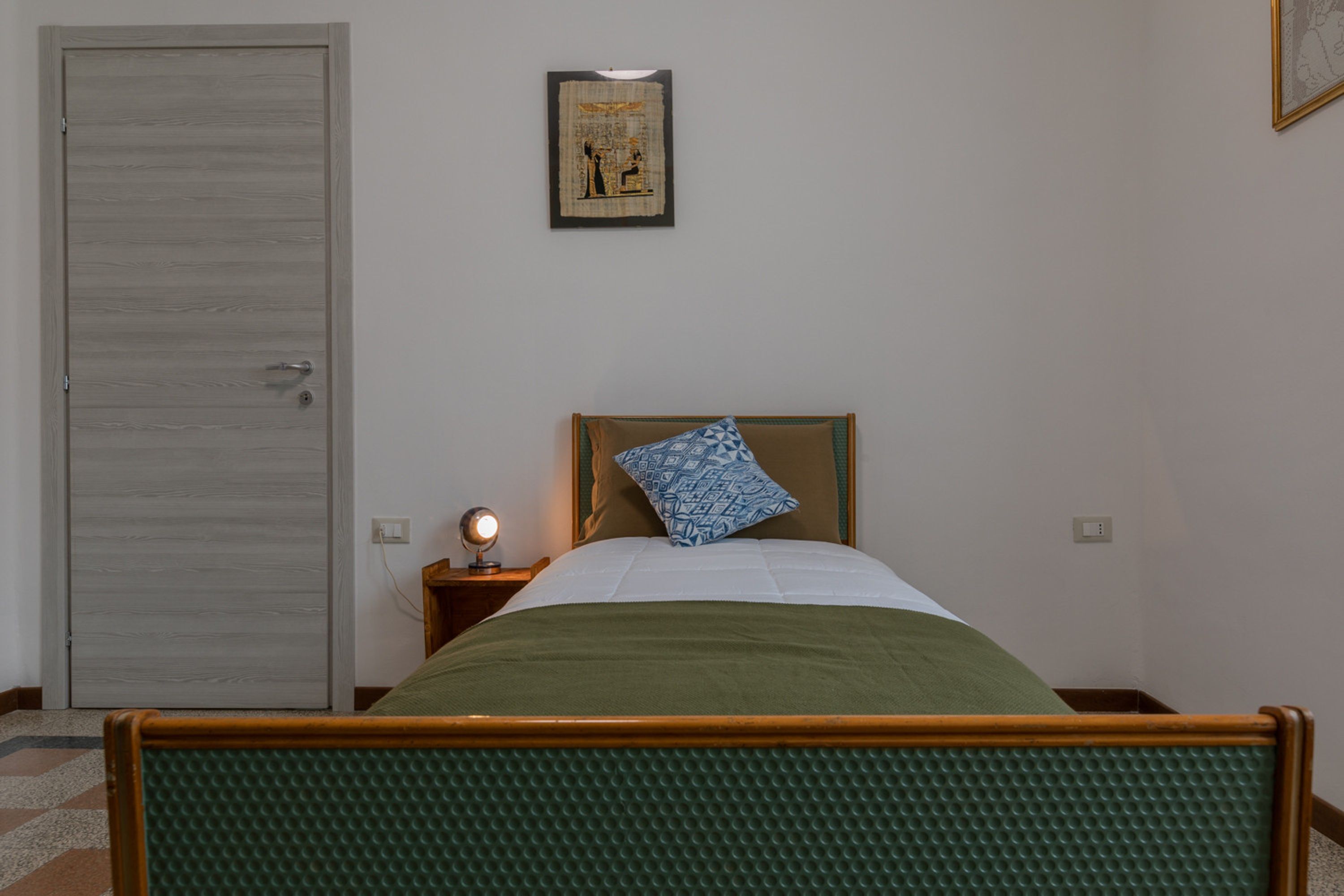 A bedroom with a wooden bed frame, a green mattress, a blue patterned pillow, a bedside table with a lamp, and a door.