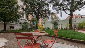 Un pati amb una taula rodona i caires vermelles, envoltat de vegetació i edificis al fons.