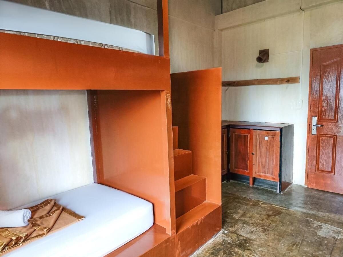 Una cama litera con un colchón blanco y una escalera de madera. Se observa una pequeña mesa de madera y una puerta al fondo.