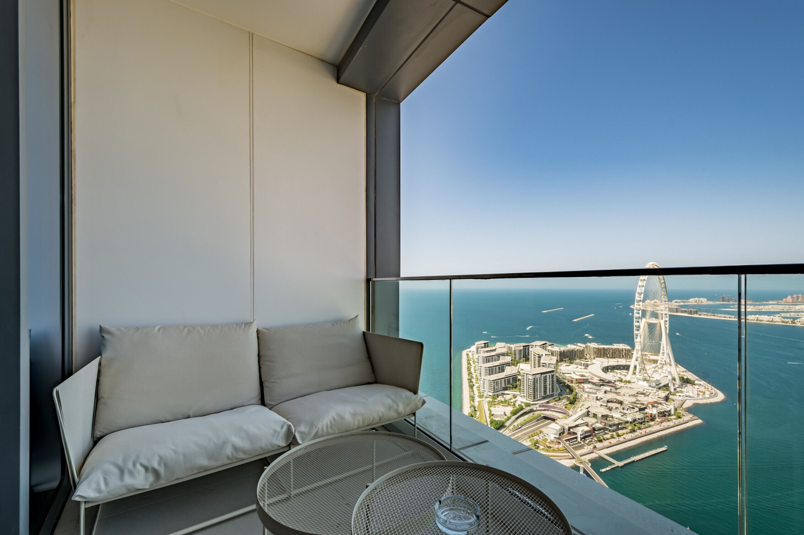Ein Balkon mit einer Couch und einem Tisch, mit Blick auf eine Küstenstadt mit modernen Gebäuden und einem markanten Riesenrad.