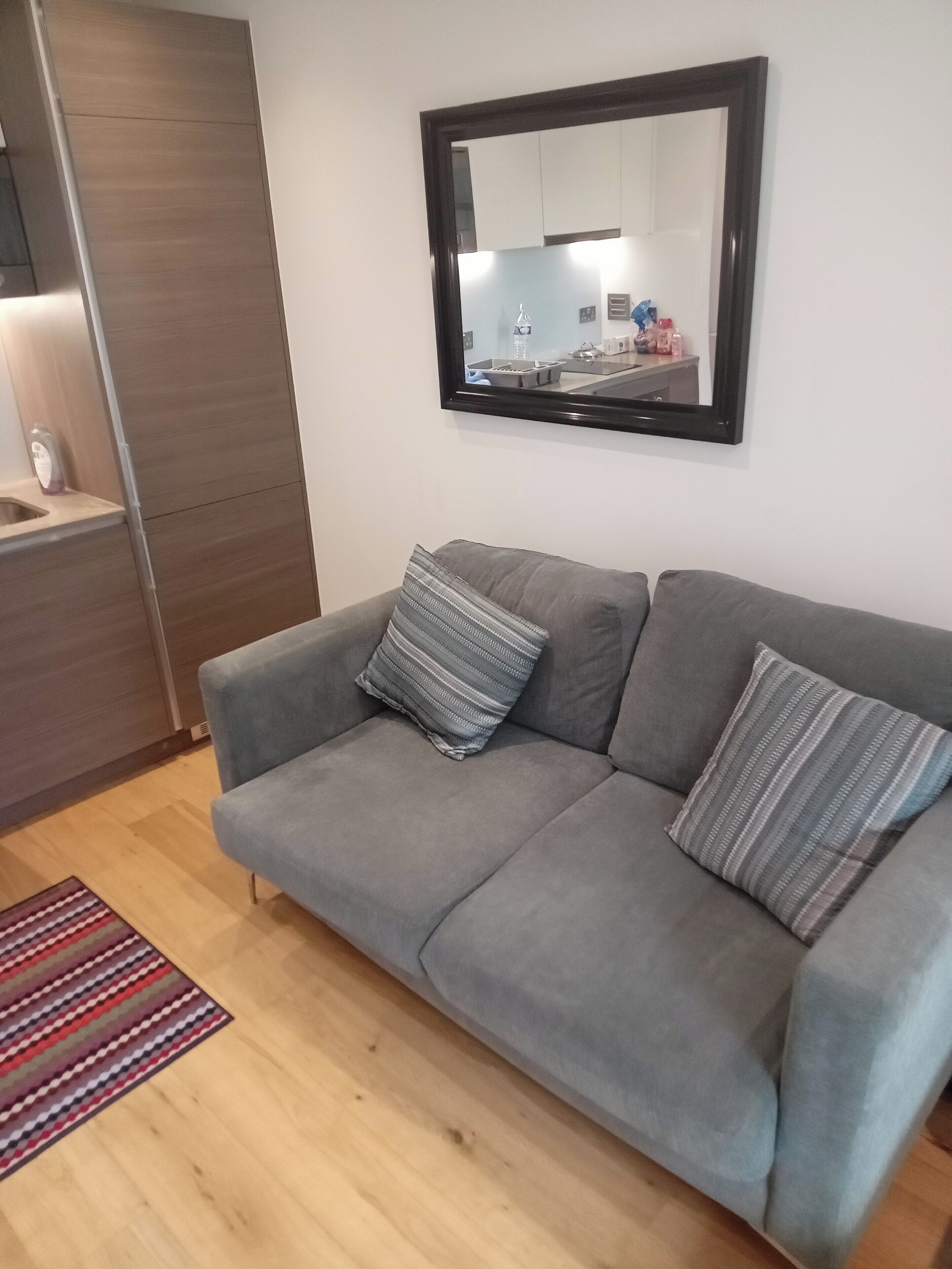 A modern living room with a grey sofa, striped cushions, a wooden floor, and a kitchen visible in the background.