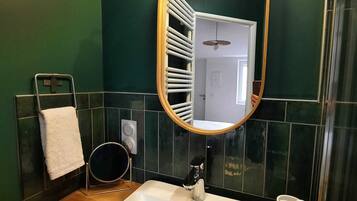 Un baño con un lavabo blanco, un espejo redondo con marco de madera, un perchero y un inodoro visible a través de un panel de vidrio.