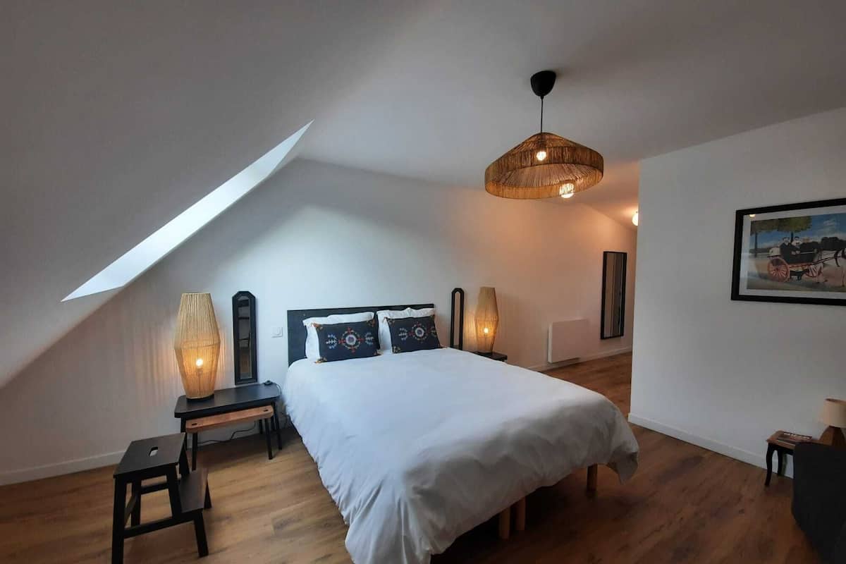 A bedroom with a sloped ceiling, a bed with white bedding, a hanging light fixture, and a framed picture on the wall.