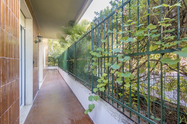Un balcón techado con piso de baldosas, cerca de metal verde y plantas trepadoras.