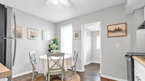 Un coin repas avec une table ronde, des chaises et un tapis tissé. On y trouve également un réfrigérateur, une cuisinière et des tableaux encadrés accrochés aux murs.