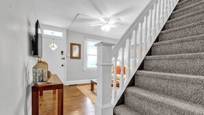 Un escalier avec une rampe blanche et une moquette grise, menant à une pièce dotée d’un plancher en bois et d’un ventilateur de plafond.