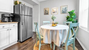 Une cuisine moderne avec un réfrigérateur en acier inoxydable, des meubles blancs et une table ronde recouverte d’une nappe blanche. On y trouve deux chaises, une machine à café et une plante en pot sur le plan de travail.