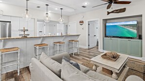 A modern kitchen with white cabinetry, a bar area with stools, a ceiling fan, and a TV displaying a coastal view.
