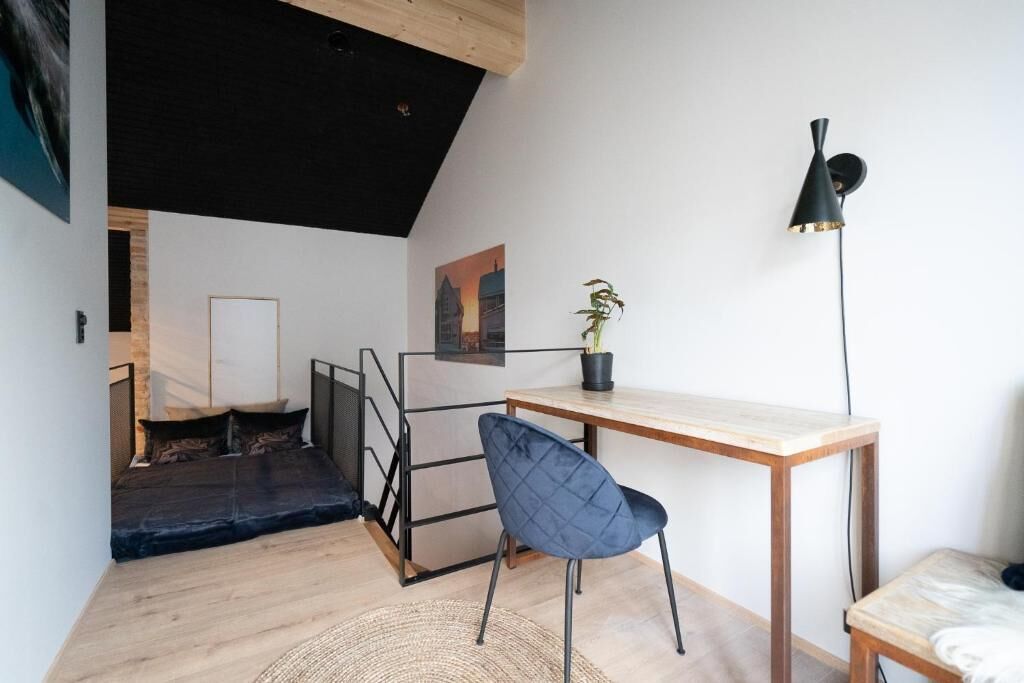 A modern loft bedroom with a sloped ceiling, a wooden desk, a blue chair, a black floor lamp, and a staircase leading to an upper level.