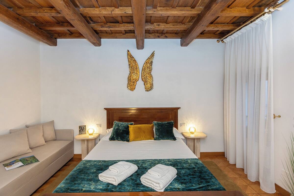 A bedroom with a wooden ceiling, a bed with white bedding, two pillows, a bedside table with a lamp, and a window with white curtains.