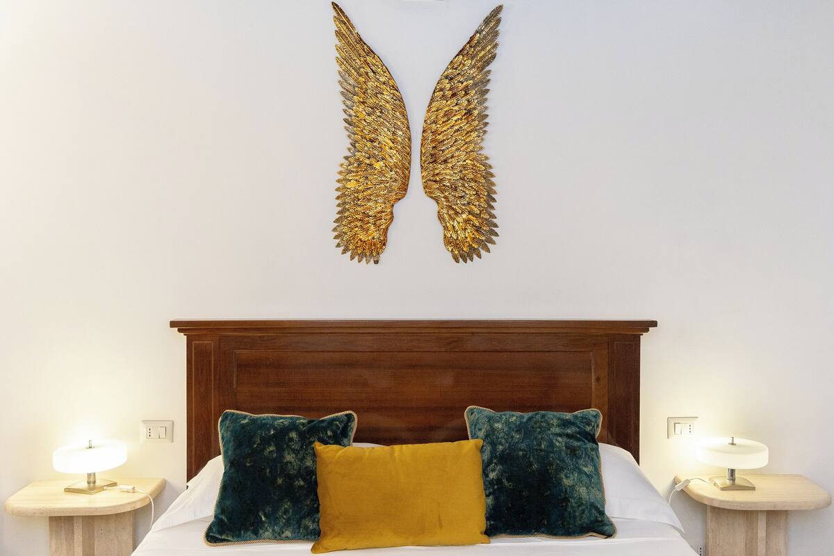 A bedroom with a wooden headboard, two bedside lamps, a bed with a mustard-colored pillow and two dark pillows, and a wall-mounted golden decorative piece.