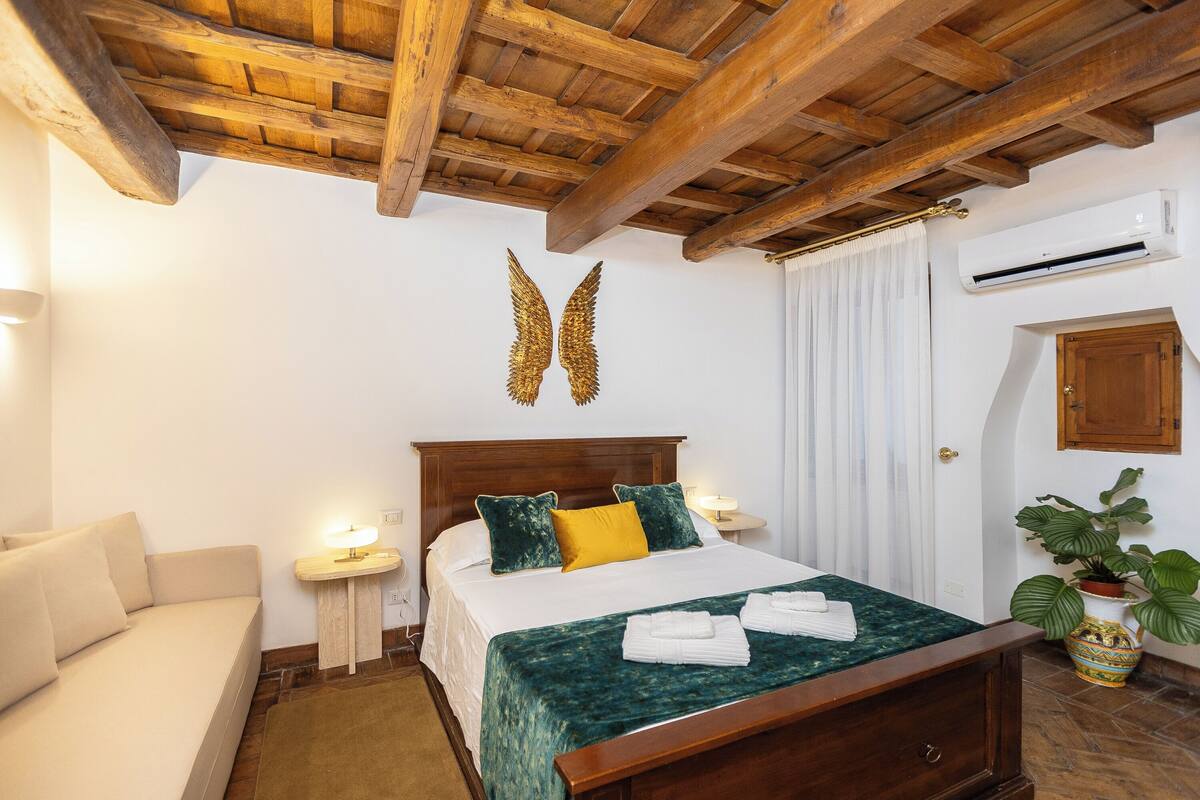 A hotel room with a wooden ceiling, a bed with a green bedspread, a bedside table with a lamp, a sofa, and a potted plant.