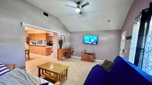 A living room with a sofa, a TV, a ceiling fan, and a kitchen in the background. - 3BR Home • Sleeps 10 • Near I-95 & Kings Bay (Kingsland)