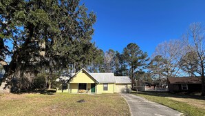 A yellow single-story house with a green door, surrounded by trees and a clear sky. - Sleeps 10•Near HWY I-95,40,17 & Kings Bay navybase,Cumberland Island,JAX airport (Kingsland)
