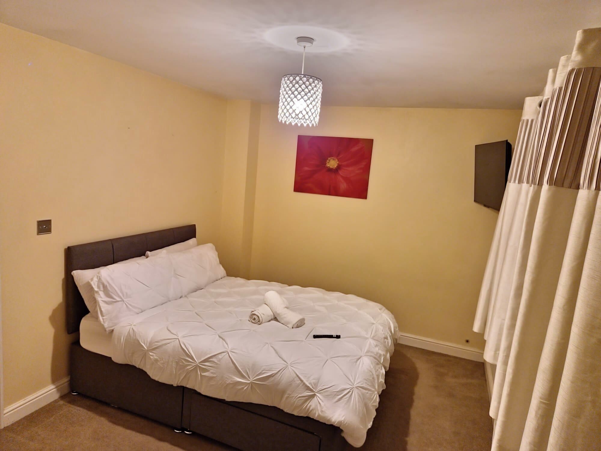 Ein ordentlich bezogenes Bett mit weißer Decke und Kissen, ein rotes Blumenbild an der Wand und eine Hängeleuchte.