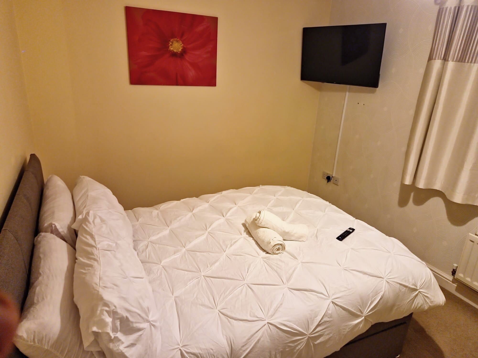A neatly made bed with a white duvet, a rolled towel, and a remote control on it. There is a red artwork on the wall and a flat-screen TV mounted above the bed.