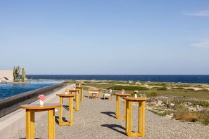 Zona de piscina amb taules i cadires de fusta, un cactus i vista a l'oceà.