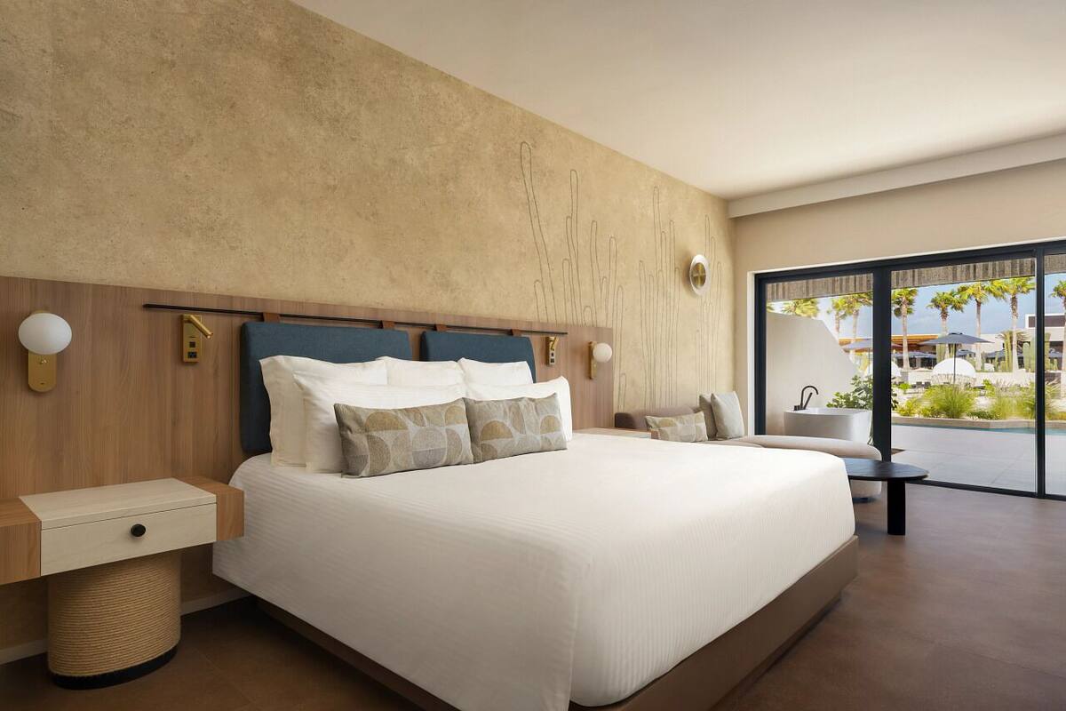 A modern hotel room with a large bed, a wooden headboard, a small bedside table, a round bedside lamp, and a view of the outdoors through large windows.