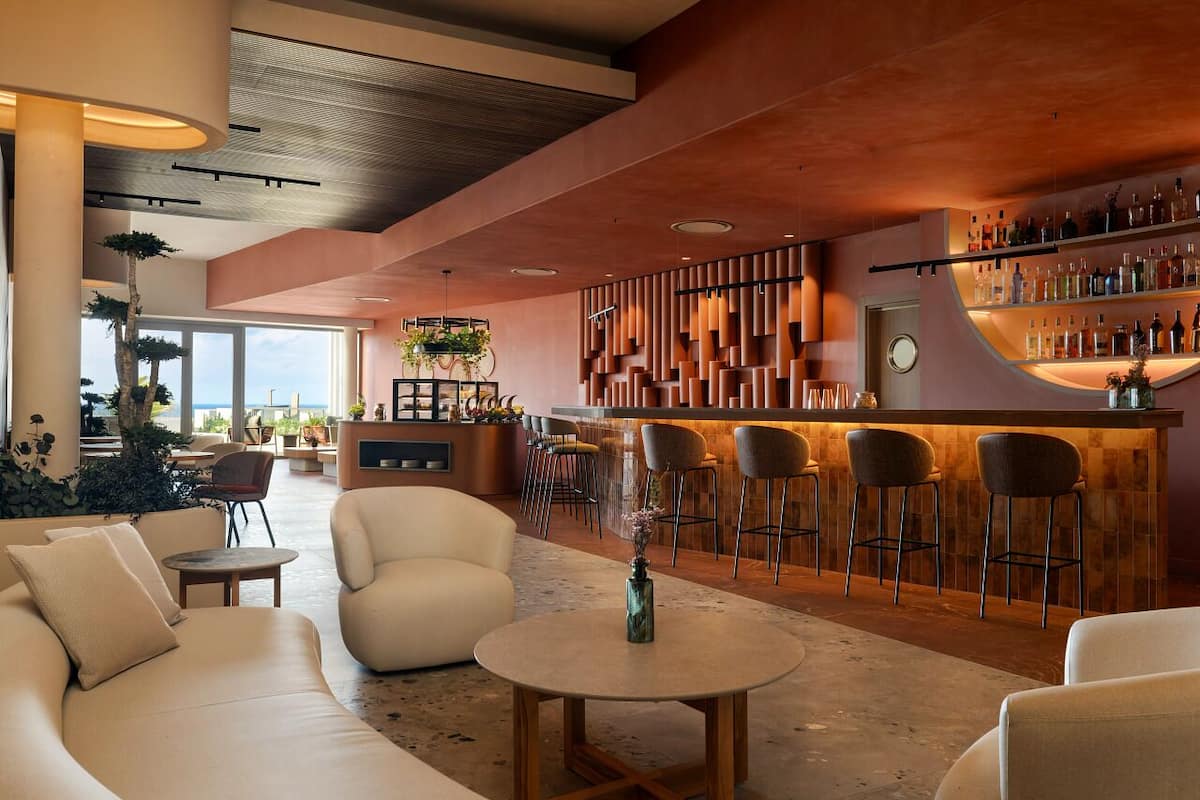 A modern bar area with a wooden bar counter, multiple stools, and a variety of bottles. There is a seating area with sofas and a round table.