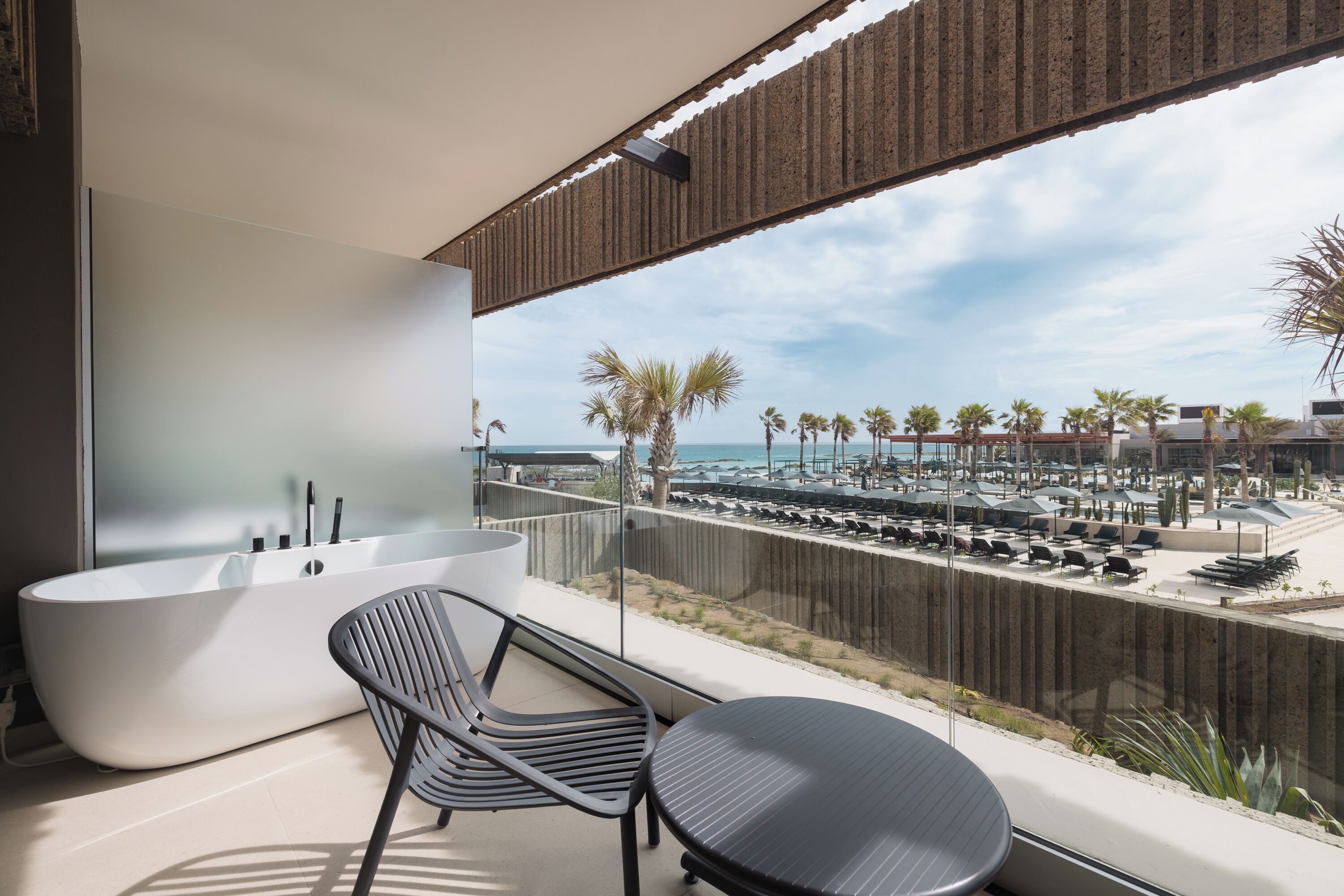 A balcony with a modern bathtub, a chair, and a table overlooking a beach with lounge chairs and palm trees.