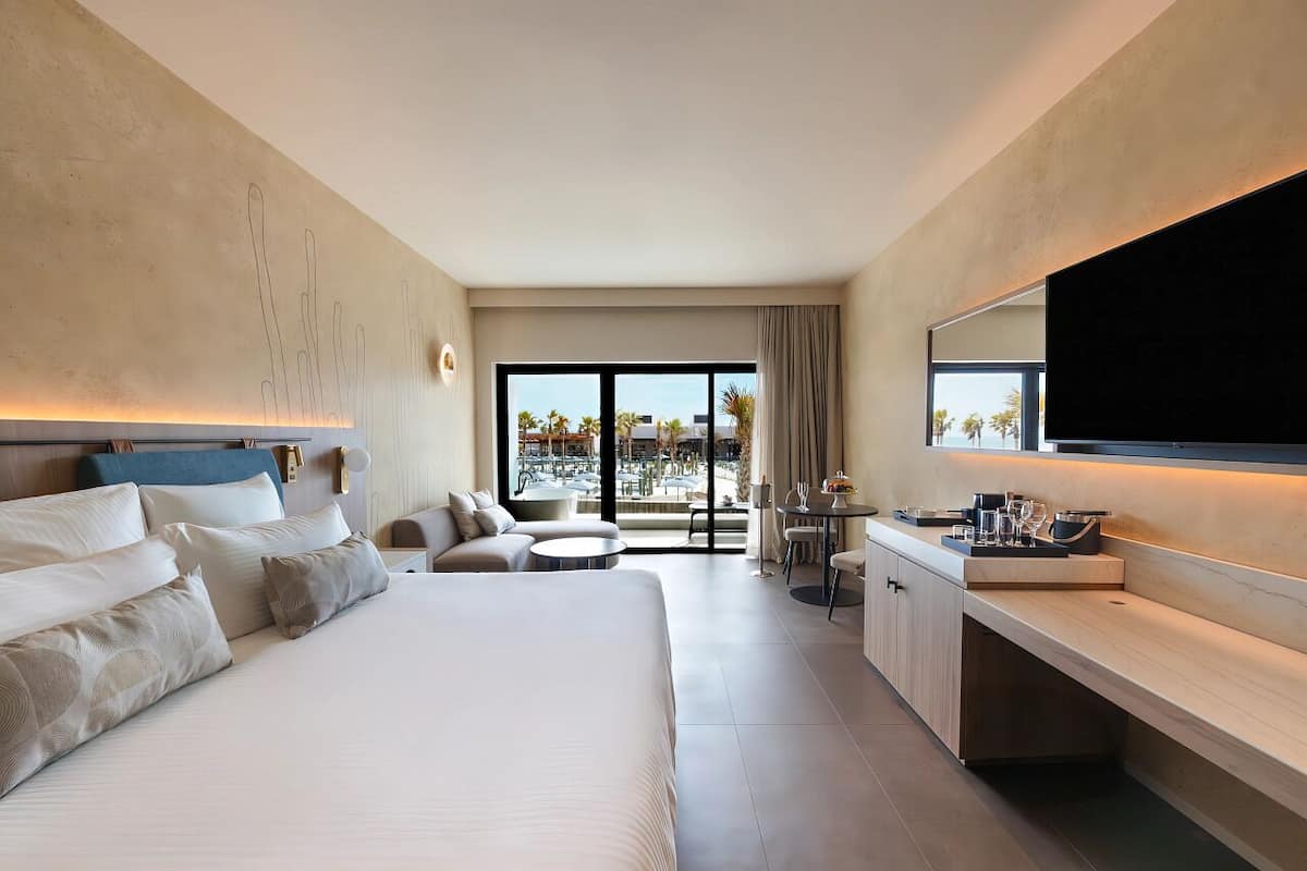 A modern hotel room with a large bed, a sitting area with a view of palm trees, a dining table, and a large mirror.