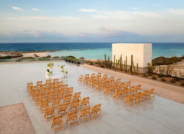 An outdoor ceremony area with wooden chairs arranged in rows, a white structure, and a view of the ocean.