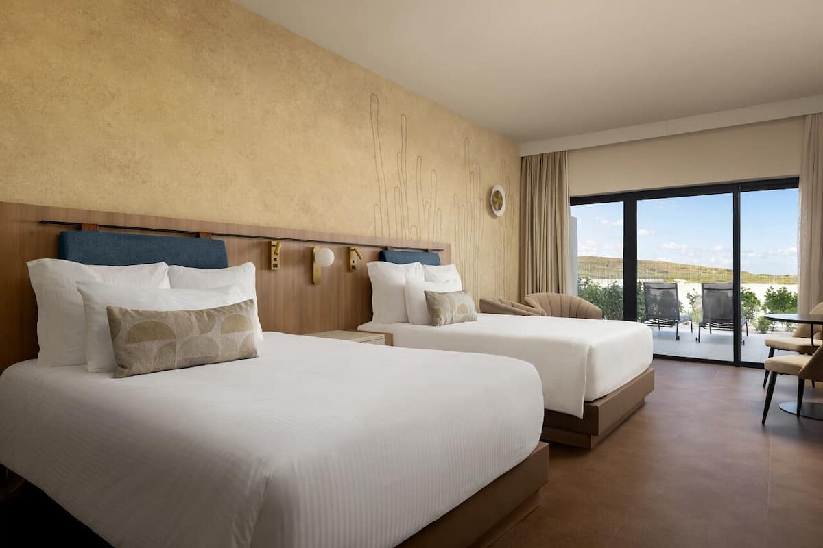 A hotel room with two beds, a large window with a view, and a balcony with outdoor furniture.
