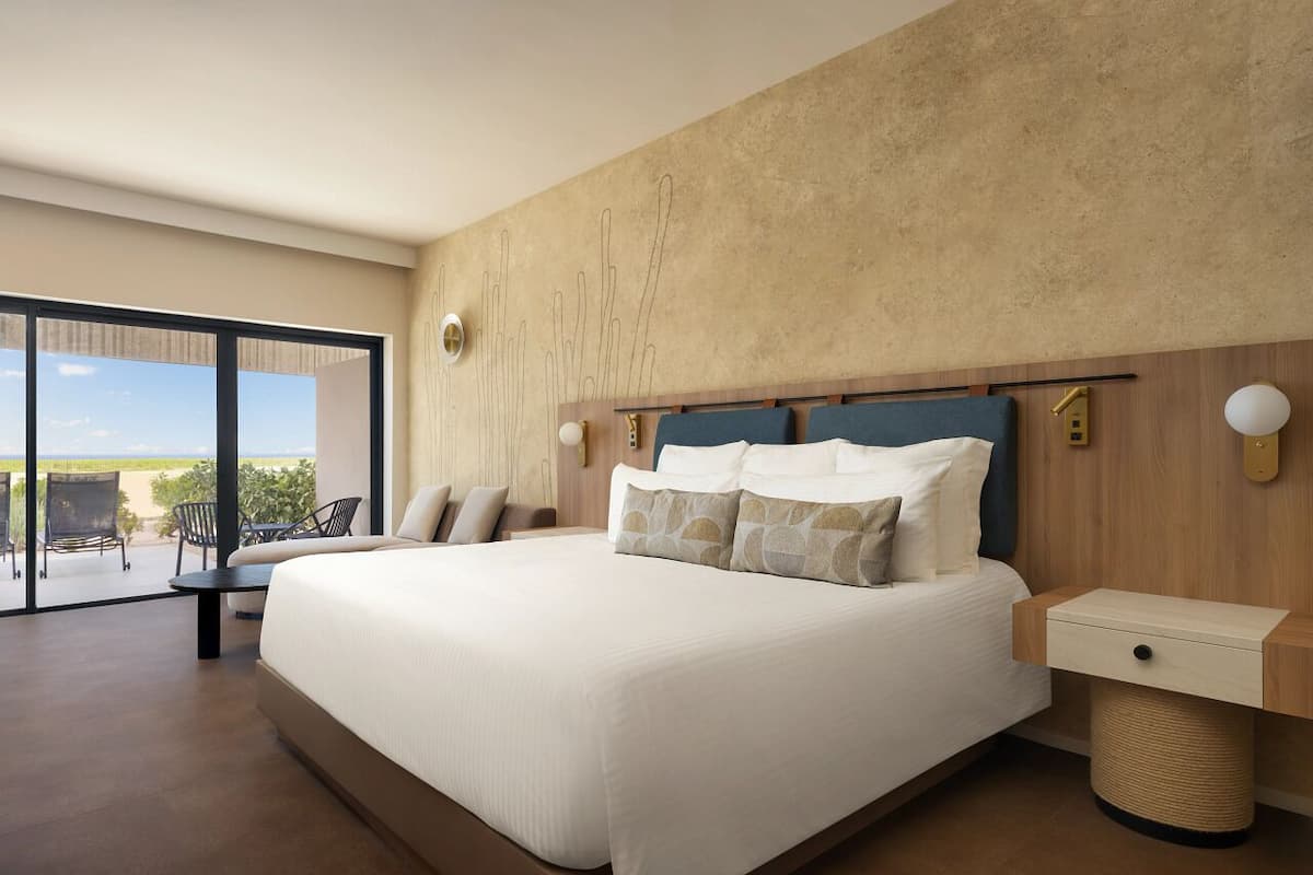 A modern bedroom with a large bed, a wooden headboard, a bedside table, and a view of the outdoors through a sliding glass door.