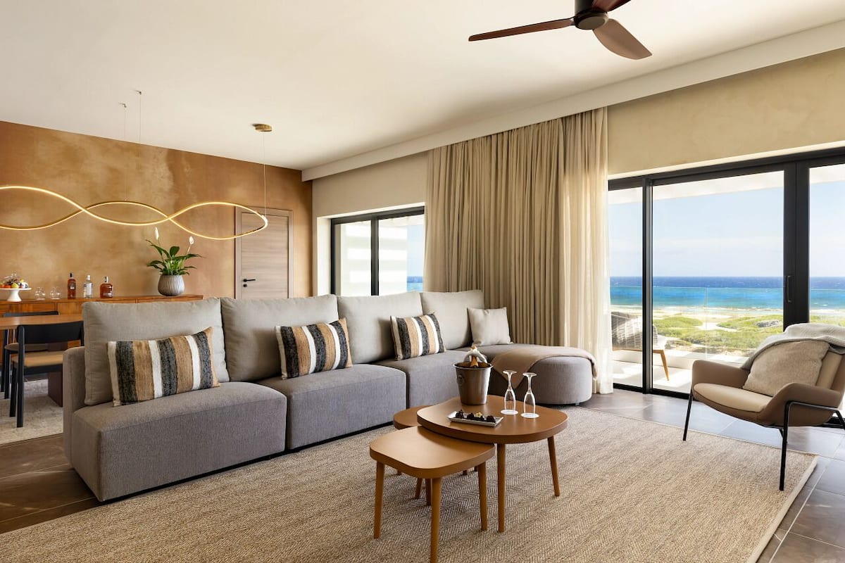 A modern living room with a large gray sofa, a wooden coffee table, and a view of the ocean through large windows.