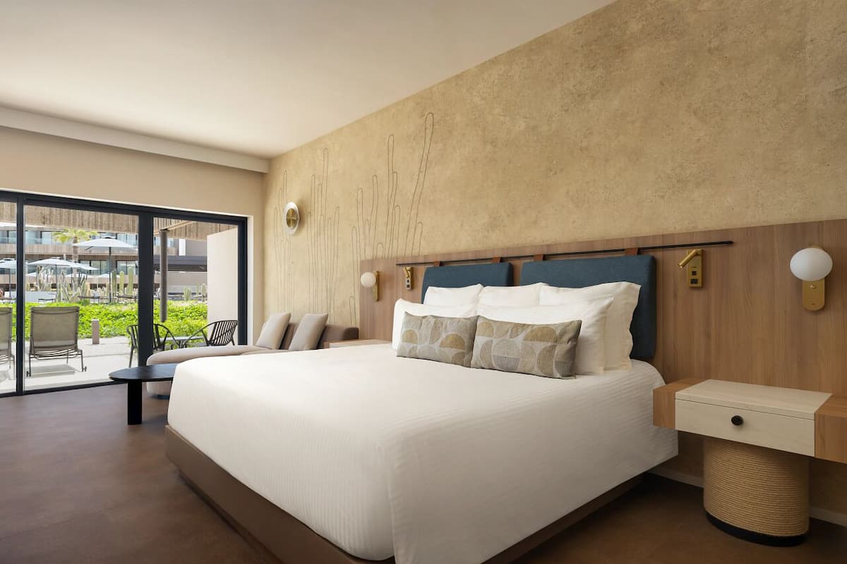 A modern hotel room with a large bed, a bedside table, a lamp, and a view of the outdoor area through large windows.