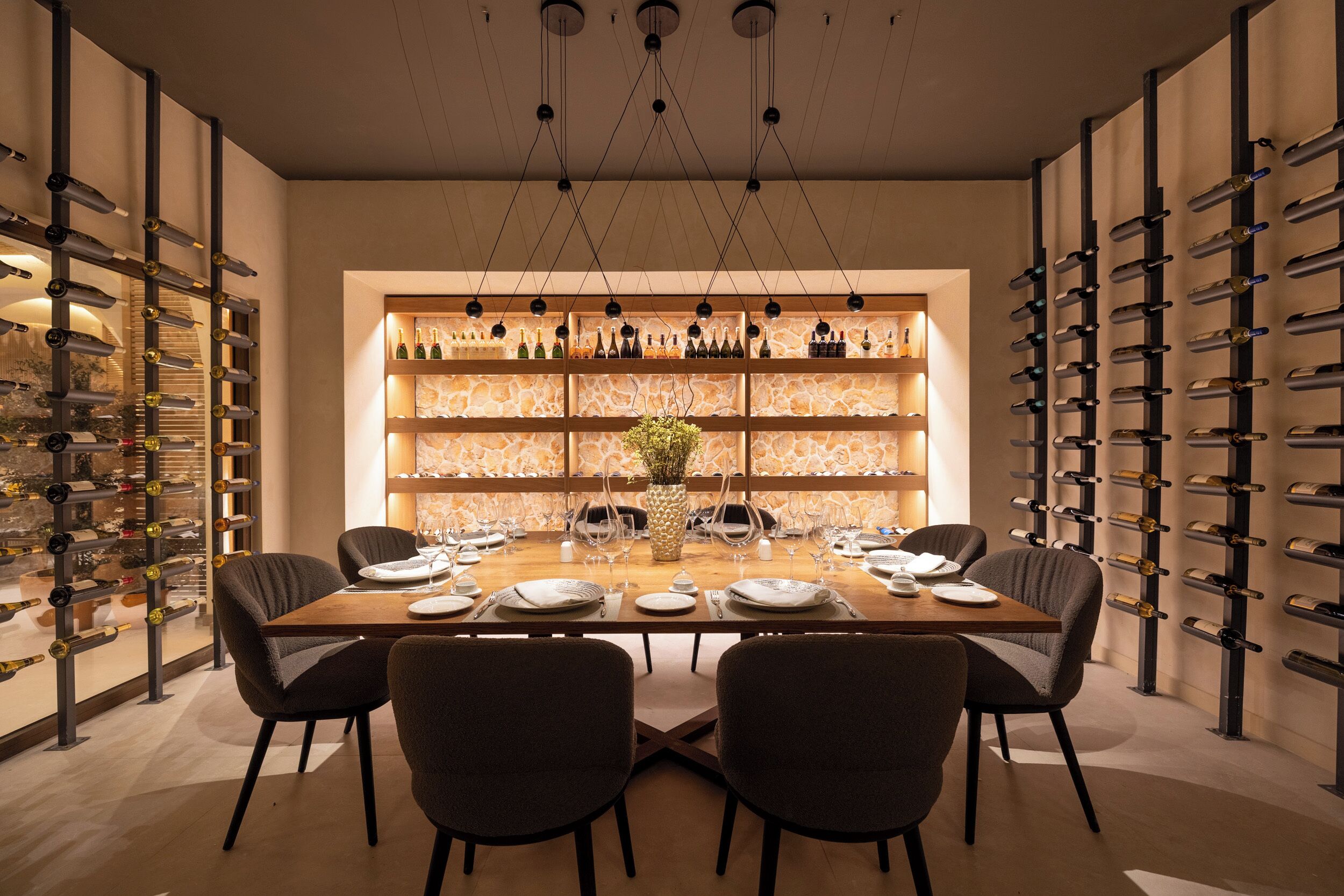 A dining area with a long table set for four, surrounded by chairs, with wine racks on both sides.