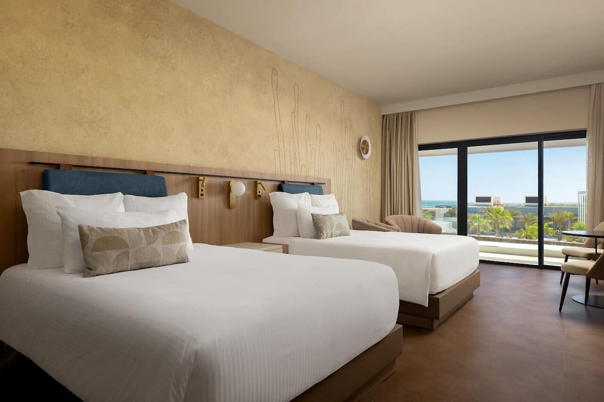 A hotel room with two beds, a large window with a city view, a small table, and a chair.