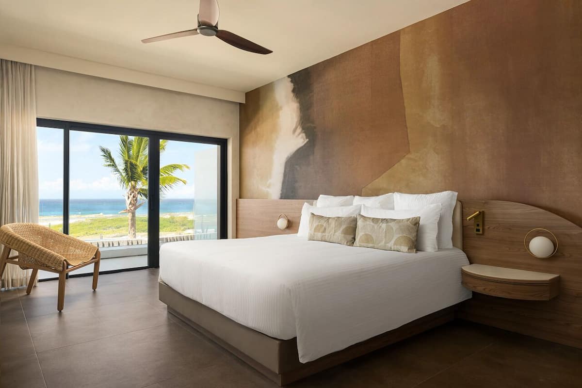 A modern hotel room with a large bed, a view of the ocean, a ceiling fan, and a large window.