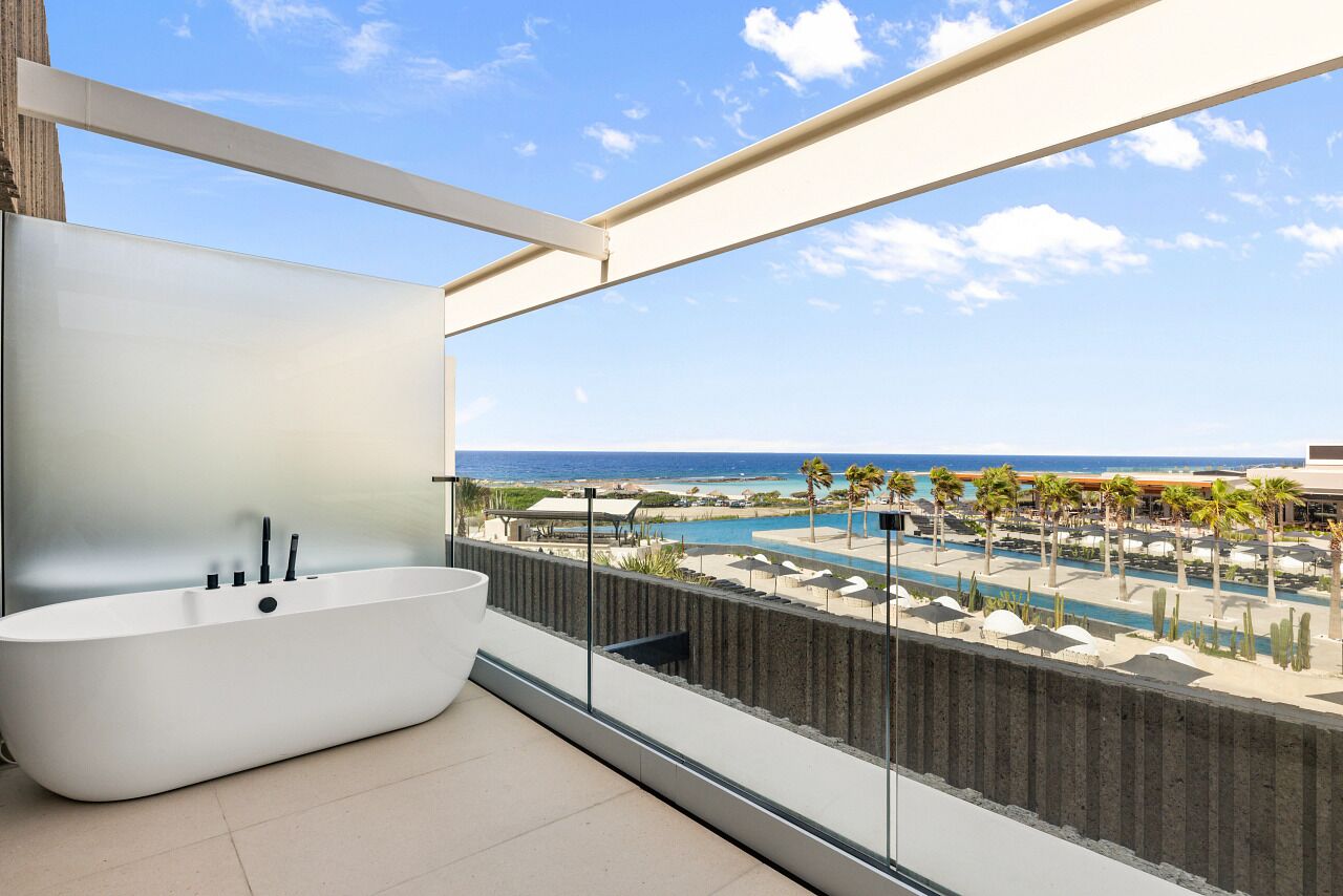 A modern bathroom with a freestanding bathtub, a large window overlooking a pool and beach area.