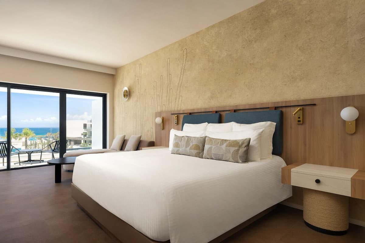 A modern hotel room with a large bed, a wooden headboard, a small bedside table, and a view of the ocean through large windows.