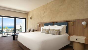 Una habitación de hotel moderna con una cama grande, un cabecero de madera, una mesita de noche pequeña y vistas al océano a través de amplias ventanas.