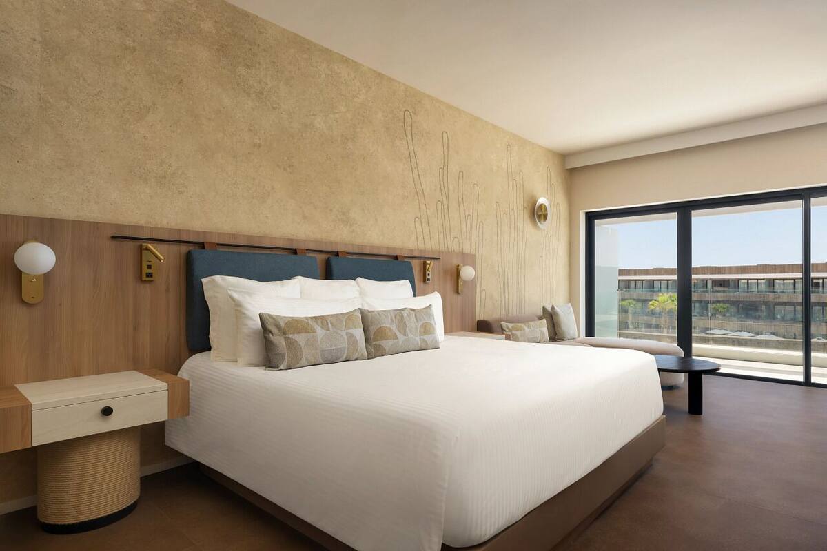 A modern hotel room with a large bed, a wooden headboard, a bedside table, a small bedside table, a round side table, and a large window with a view of the city.