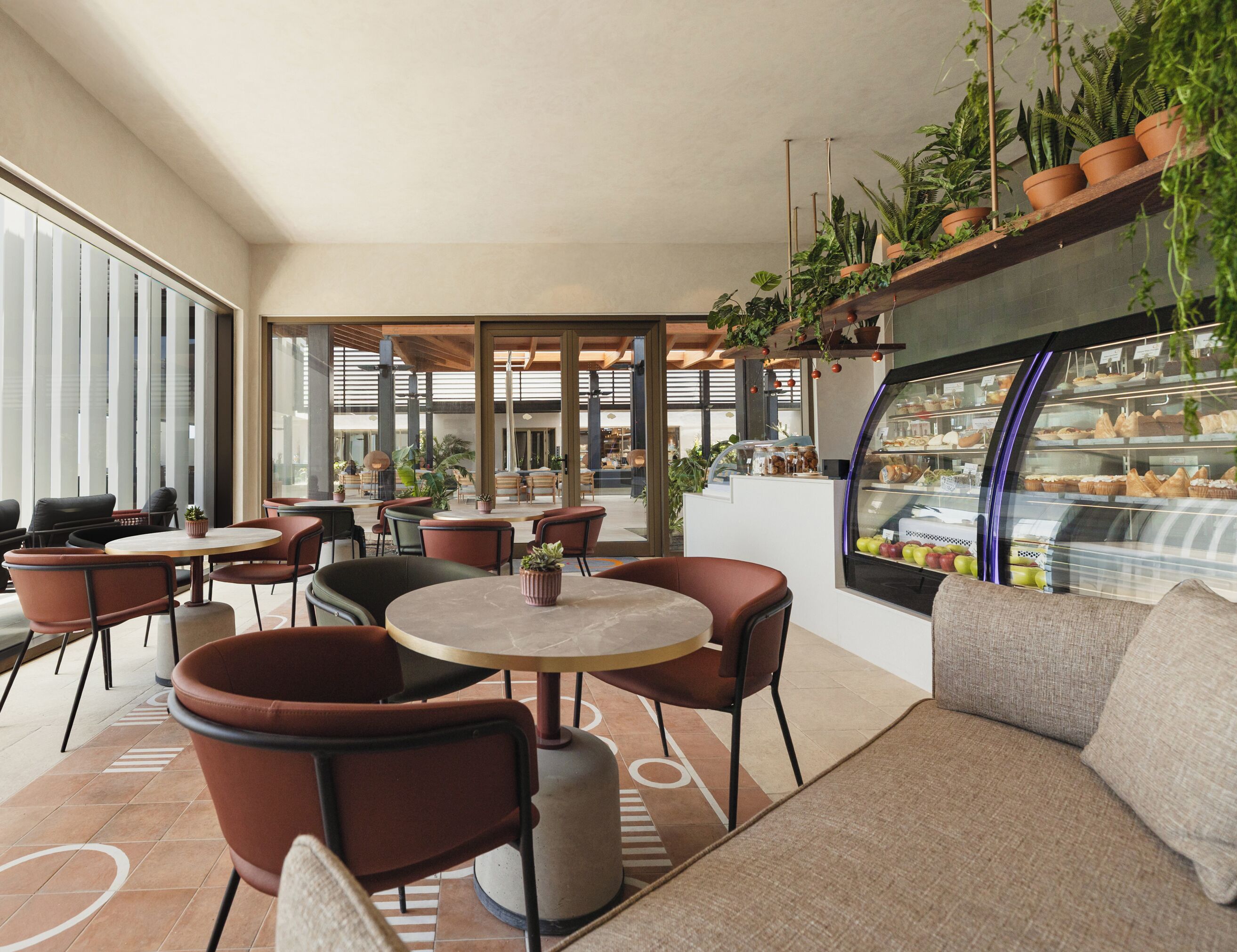 Un café moderno con mesas y sillas redondas, una vitrina de panadería y plantas en macetas.