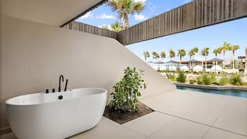 Un baño moderno con una bañera independiente, una pequeña planta y vistas a una piscina con palmeras y sombrillas.