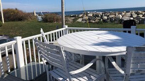 A deck with a round table and white chairs, overlooking a rocky shoreline with a boat in the distance. - Oceanfront 1-bedroom condo with Deck Views in Rye (Rye)