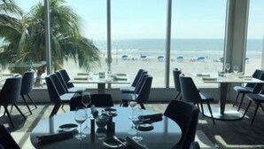 A dining area with tables and chairs, large windows with a beach view, and palm trees outside. - Ocean/Pool View 703- Relax and recharge at Fort Myers Beach (Fort Myers Beach)