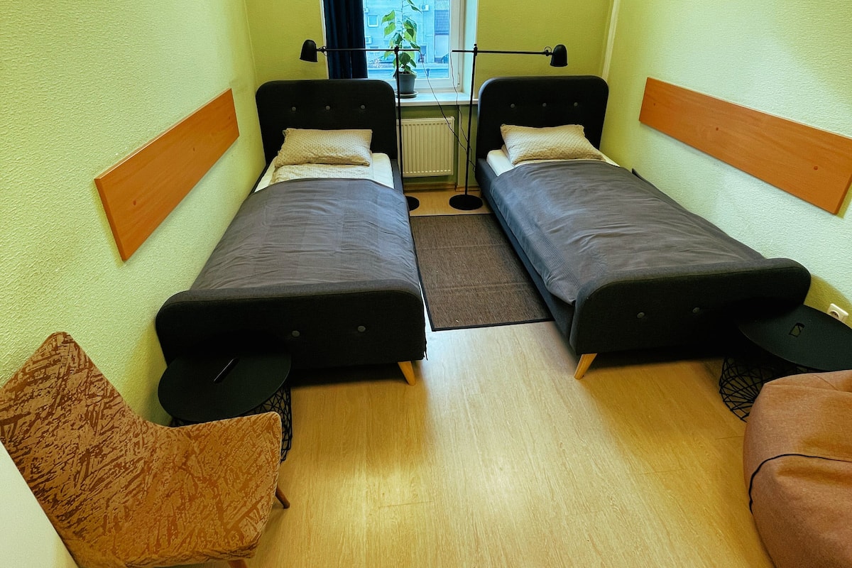 A small, compact room with two single beds, a small table, a chair, and a radiator.