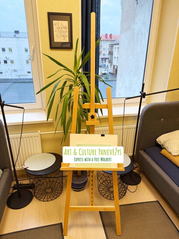 A room with a wooden easel, a potted plant, a framed picture on the wall, and a view of buildings through a window. - Art & Culture Panevėžys, Self Check-In (Panevezys)