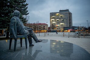 A public square with a statue of two seated figures, a modern building with illuminated signage, and a clear sky. - Art & Culture Panevėžys, Self Check-In (Panevezys)