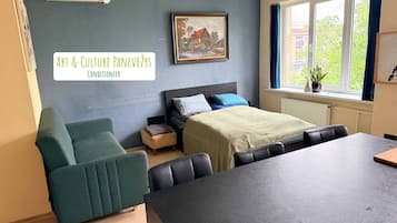 A room with a bed, a table, a chair, a small stool, and a painting on the wall.