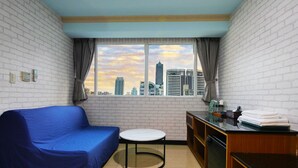 A room with a blue sofa, a small round table, a kitchen area with a counter and cabinets, and a window with a cityscape view. - IHI loveriver Hotel (Kaohsiung)